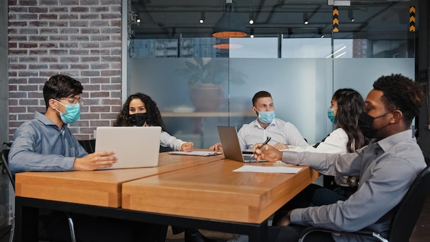 Verschiedene Geschäftsteams, Mitarbeiter in medizinischen Masken, die im Büro zusammenarbeiten, gemischtrassige Kollegen, multiethnische Gruppenteamarbeit, die Projektarbeit im Konferenzraum diskutieren, überprüfen Papiere online