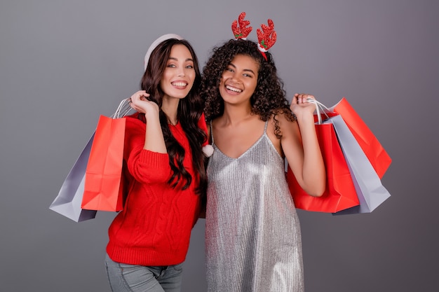 Verschiedene glückliche Frauen mit den bunten Einkaufstaschen, die den roten Weihnachtshut lokalisiert über Grau tragen