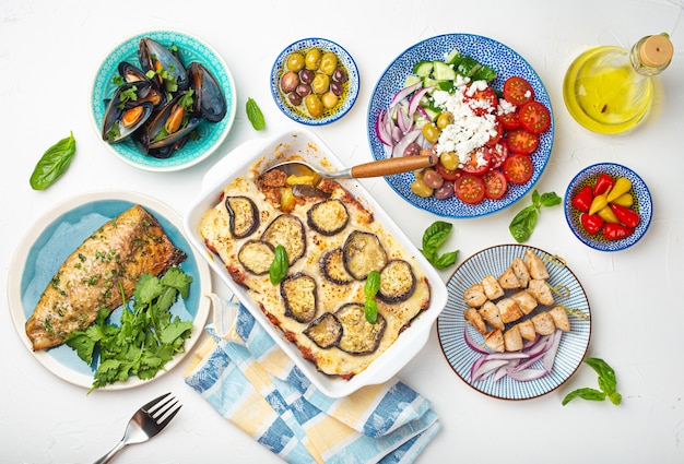 Verschiedene griechische Gerichte auf weißem Steinhintergrund von oben, Moussaka, gegrillter Fisch, Souvlaki, griechischer Salat, gedämpfte Muscheln mit Kräutern, Vorspeisen aus Griechenland von oben