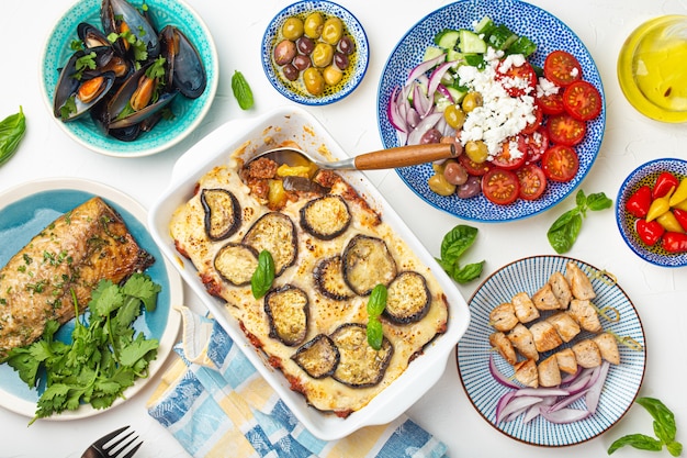 Verschiedene griechische Gerichte auf weißem Steinhintergrund von oben, Moussaka, gegrillter Fisch, Souvlaki, griechischer Salat, gedämpfte Muscheln mit Kräutern, Vorspeisen aus Griechenland von oben