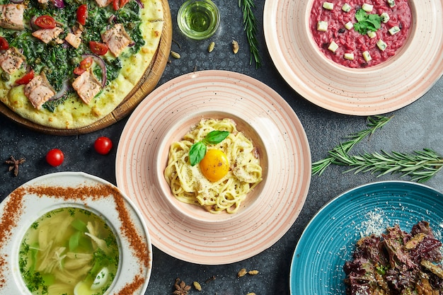 Verschiedene italienische Gerichte auf einer dunklen Oberfläche: Carbonara-Nudeln, Lachspizza, Rote-Bete-Risotto, hausgemachte Hühnerbrühe und Salat. Draufsicht Wohnung lag Essen