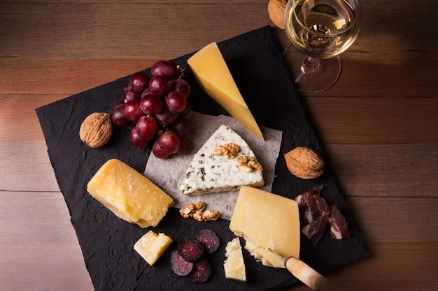 Verschiedene Käsesorten, Nüsse, Trauben, Obst, geräuchertes Fleisch und ein Glas Wein auf einem Serviertisch. Dunkler und launischer Stil. Freier Platz für Text.