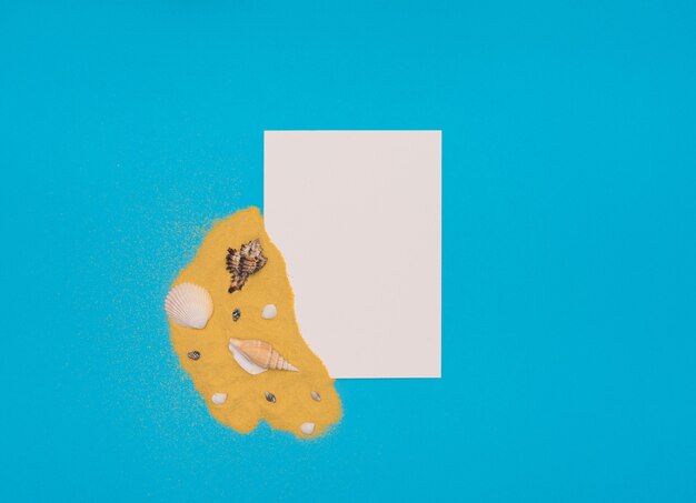 Verschiedene Muscheln auf leuchtend gelbem Sand mit weißer Papierkarte auf blauem Hintergrund Sommerzeit