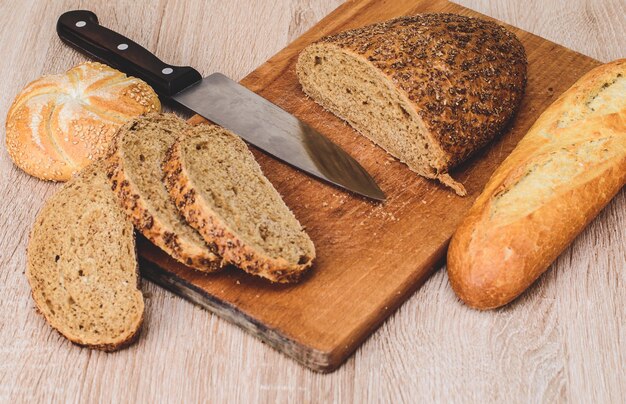 Verschiedene Rassen auf hölzernem Hintergrund. Messer auf einem Holzbrett mit Brot und verschiedenen Brötchen auf dem Tisch. Knuspriges Brot auf einem Holzbrett.