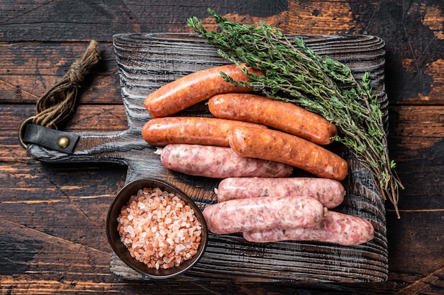 Verschiedene rohe Schweine- und Rindfleischwürste mit Gewürzen auf einem Holzbrett mit Thymian. Dunkler hölzerner Hintergrund. Ansicht von oben.