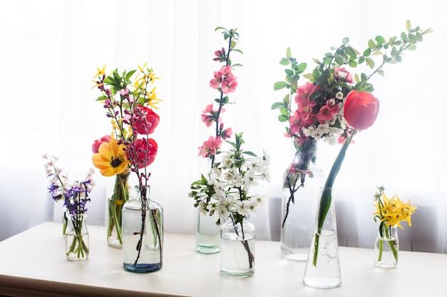 Verschiedene schöne Blumen in Gläsern mit Wasser auf dem Tisch am Fenster