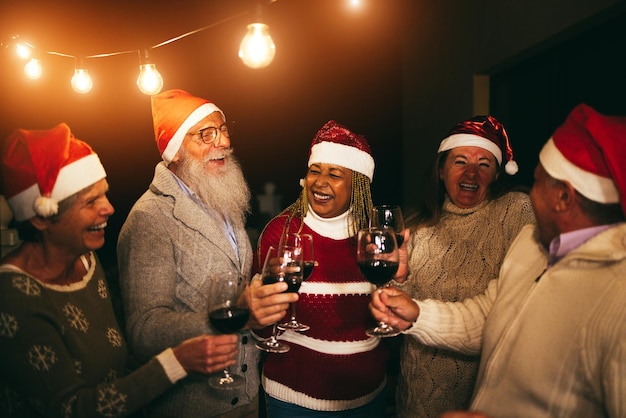 Verschiedene Senioren haben Spaß daran, an Heiligabend Wein zu trinken und Weihnachtsmannmützen zu tragen. Weicher Fokus auf das Gesicht einer afrikanischen Frau