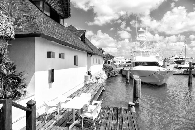 Verschiedene Yachten in der Dominikanischen Republik auf dem Wasser in der Bucht in der Nähe des weißen Gebäudes mit Tisch und Stühlen am sonnigen Tag mit Wolken am blauen Himmel