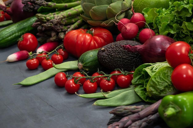 Verschiedenes Bio-Gemüse - Spargel, Tomatenkirsche, Avocado, Artischocke, Pfeffer, Limette, Rötlich, Blumenkohl, grüne Bohnen, Gurke auf grauem Hintergrund.