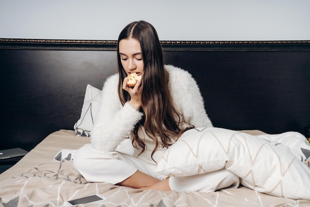 Verschlafenes langhaariges Mädchen im weißen Pyjama wacht morgens früh auf, sitzt auf dem Bett und isst einen Apfel zum Frühstück
