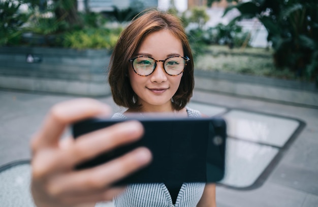 Verschwommene lächelnde erwachsene schöne asiatische Frau in lässigem Outfit und Brille, die ein Selfie-Foto macht