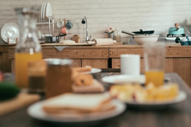 Verschwommene Sicht auf eine gesunde Mahlzeit auf dem Küchentisch Frischer Orangensaft, Toastbrot und Marmelade am Morgen zu Hause. Holzkochstelle im Hintergrund mit handgemachtem Teig un Backcroissant mit Nudelholz.