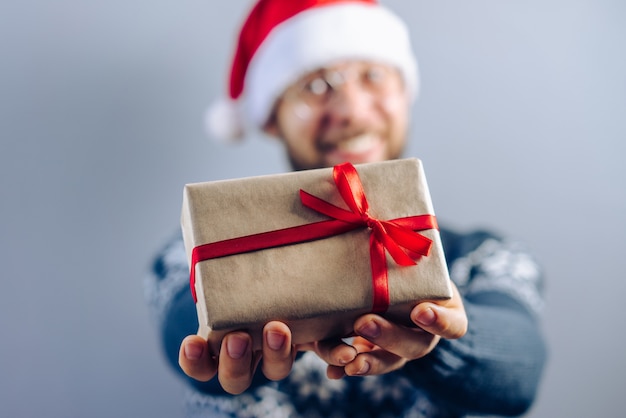 Verschwommenes Nahaufnahmebild eines bärtigen Kerls, der Weihnachtsmann-Hut und Brillen trägt, die ein Geschenk teilen, das in Bastelpapier eingewickelt und mit satinrotem Band verziert wird