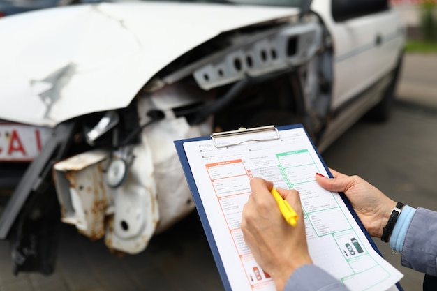 Versicherungsvertreter hält Zwischenablage und Kugelschreiber-Nahaufnahme und zerstörtes Auto im Hintergrund