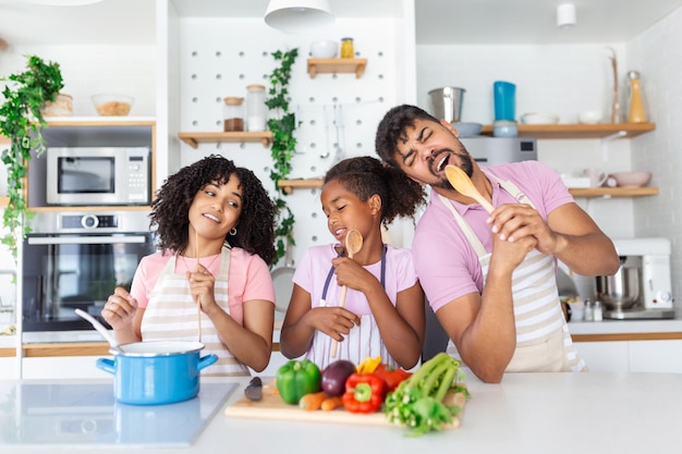 Verspielte afroamerikanische Familie, die Spaß in der Küche hat, singt und tanzt, während sie das Mittagessen zu Hause kocht Fröhliche Familie mit Tochter, die Spachtel als Mikrofone verwendet Freiraum