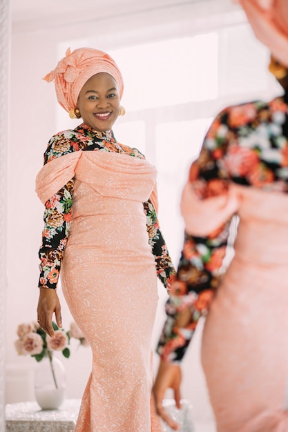 Verspielte, charmante afrikanische Dame in einem eleganten, trendigen rosa Kleid mit ethnischem Aufdruck, traditionellem Kopftuch, die in die Kamera lächelt, während sie ihr Spiegelbild betrachtet.