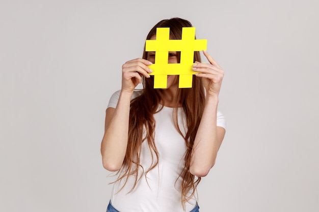 Verspielte Frau, die sich hinter gelbem Hashtag-Symbol versteckt, Gesicht mit Hash-Zeichen bedeckt, Konzept des Bloggens und virale Themen im Internet, trägt weißes T-Shirt. Studioaufnahme im Innenbereich isoliert auf grauem Hintergrund