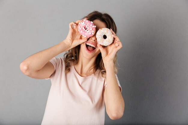 Verspielte Frau im T-Shirt, die Spaß hat, während Donuts auf ihren Augen über Grau halten