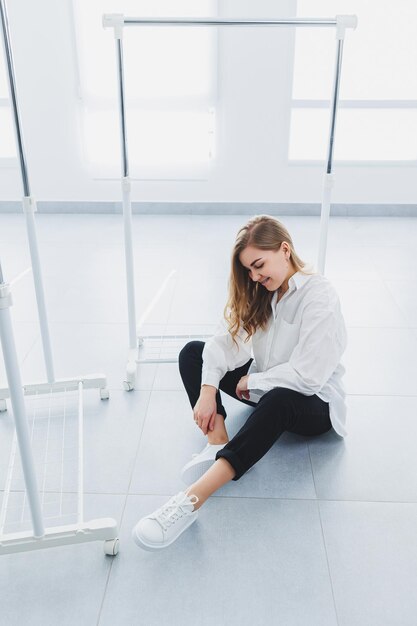 Verspielte, selbstbewusste Frau in einem stylischen weißen Baumwollhemd, schwarzen Jeans, weißen Lederturnschuhen, lässiger Stil. Blonde Frau auf grauem Hintergrund in stilvoller Kleidung