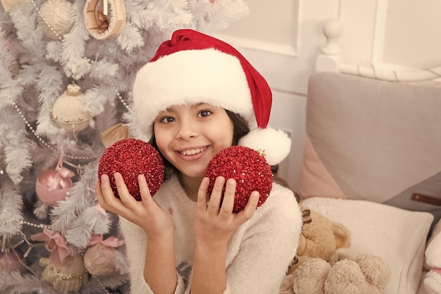 Verspielte Stimmung Festliche Atmosphäre schaffen Kind schmücken Weihnachtsbaum mit roter Kugel Mädchen Kind schmücken Weihnachtsbaum Kind in Weihnachtsmütze schmücken Weihnachtsbaum Gute Stimmung Fröhliches Kind