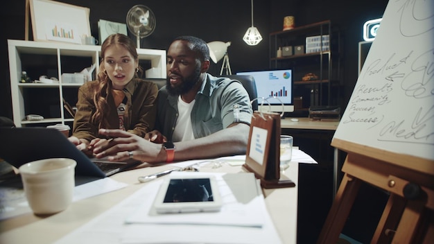 Verspielter Afro-Mann und Hipster-Frau unterhalten sich am Arbeitsplatz auf dem Laptop-Bildschirm über Daten. Geschäftspaar bespricht Projekt auf dem Bildschirm im dunklen Büro. Team gemischter Rassen arbeitet drinnen am Laptop