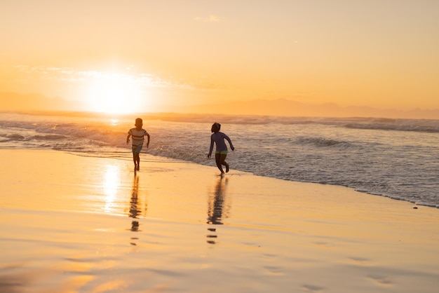 Verspielter afroamerikanischer Bruder und Schwester, die bei Sonnenuntergang am Strand gegen den Himmel laufen