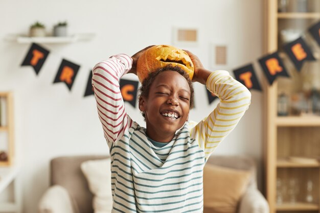 Verspielter afroamerikanischer Junge, der Spaß mit Jack O 'Lantern Kürbis hat, der ihn auf seinen Kopf setzt, mittlere Porträtaufnahme