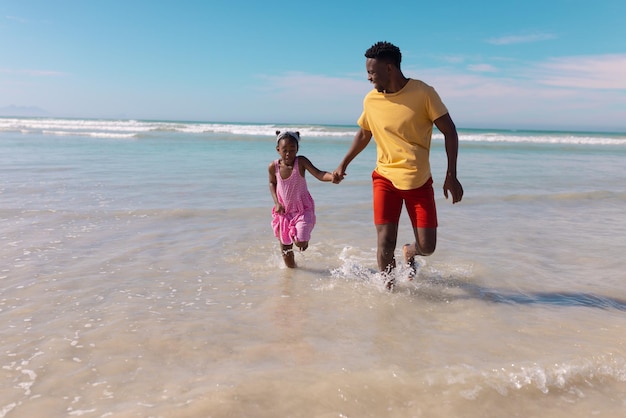 Verspielter afroamerikanischer junger Vater und Tochter halten Händchen und laufen im Meer gegen den Himmel