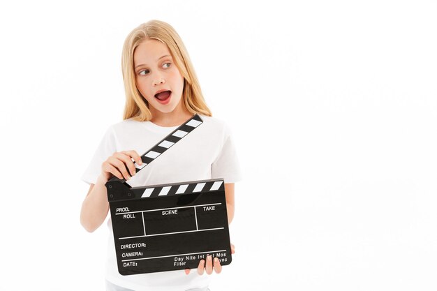 Verspieltes junges blondes Mädchen in der Freizeitkleidung, die Filmschindel hält, während weg auf Weiß schaut