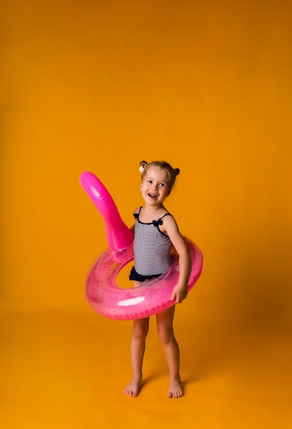 Verspieltes kleines Mädchen in einem gestreiften Badeanzug hält einen rosa aufblasbaren Kreis mit einem Flamingo auf einer gelben Oberfläche mit einer Kopie des Raumes