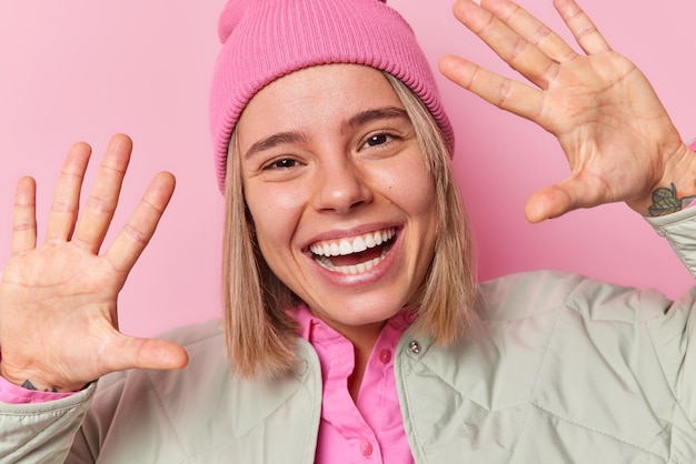 Verspieltes, positives, hellhaariges Teenager-Mädchen hält die Handflächen hoch, hat gute Laune, trägt Hut und Jacke, die in guter Laune sind, versucht, Sie zu amüsieren, posiert vor rosafarbenem Hintergrund Menschen und fröhliches Gefühlskonzept