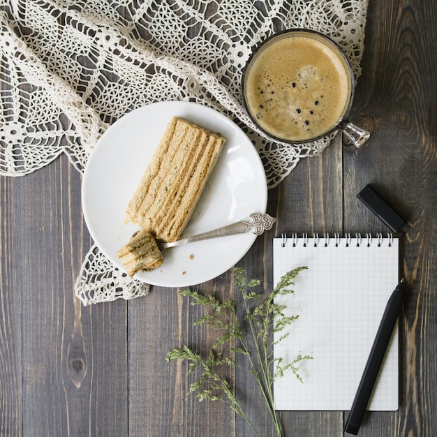 Verspotten Sie herauf Arbeitsplatz mit wildem Gras, Stift, Notizbuch, Scheibe des Kuchens und Tasse Kaffee auf hölzernem Hintergrund. Flache Lage, Draufsicht. stilvolles weibliches Konzept