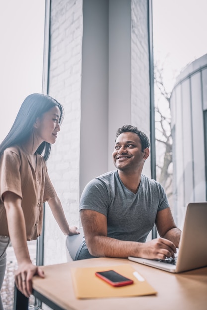 Verstehen. Lächelnder, freundlicher, dunkelhäutiger Typ, der am Laptop sitzt, und ein langhaariges asiatisches Mädchen, das in der Nähe steht und sich tagsüber im Büro ansieht