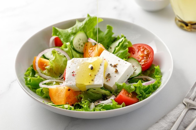 Verter aceite de oliva en ensalada griega con hojas verdes tomates pepino queso feta Tazón blanco mármol