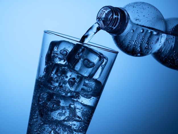 Verter agua de una botella de plástico en un vaso con hielo sobre un fondo azul claro