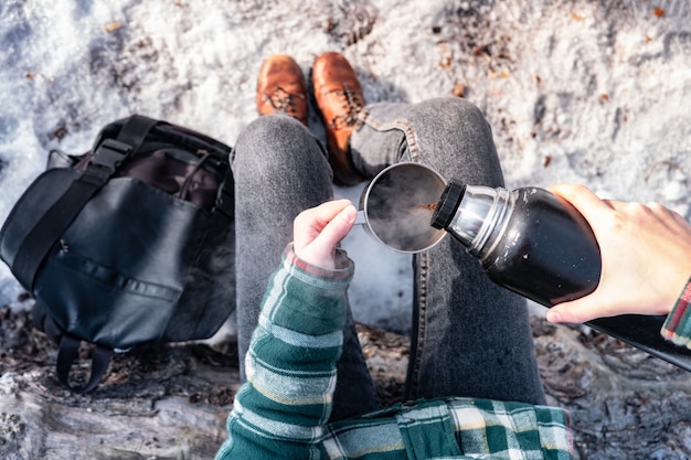 Verter una bebida caliente del termo en un campamento. Persona en un bosque de invierno durante un viaje de senderismo calentándose, toma de punto de vista