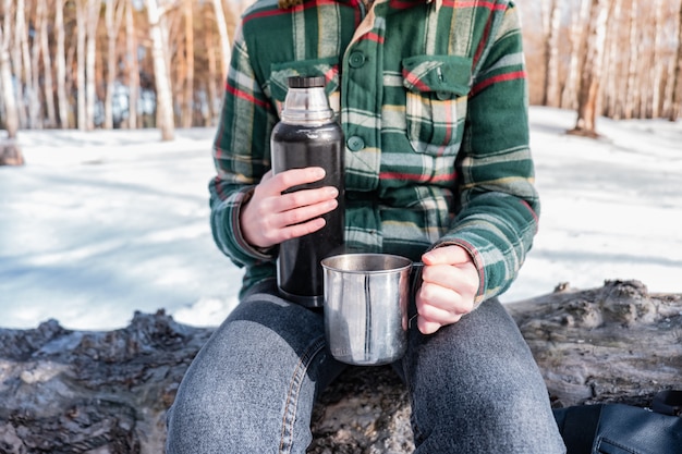 Verter una bebida caliente del termo en un campamento. Persona en un bosque de invierno durante un viaje de senderismo calentándose