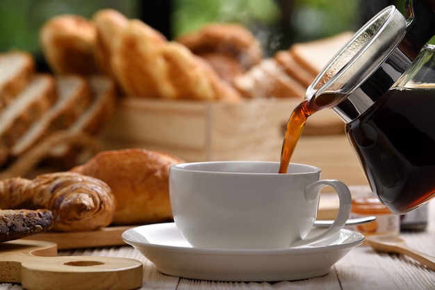 Verter café con humo en una taza con pan o bollo, croissant y panadería en mesa de madera blanca.