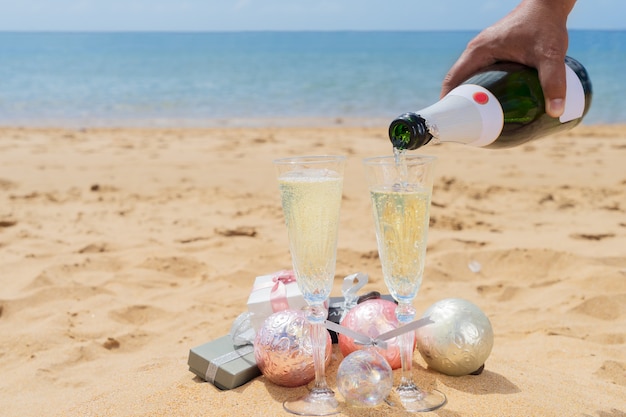 Verter champán en copas con caja de regalo de navidad y adornos en la playa junto al mar tropical