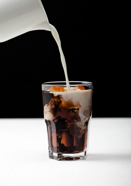 Verter la leche fresca en un vaso de café negro con cubitos de hielo en la pizarra y fondo negro.