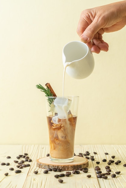 Verter la leche en un vaso de café negro con cubitos de hielo, canela y romero en la mesa de madera