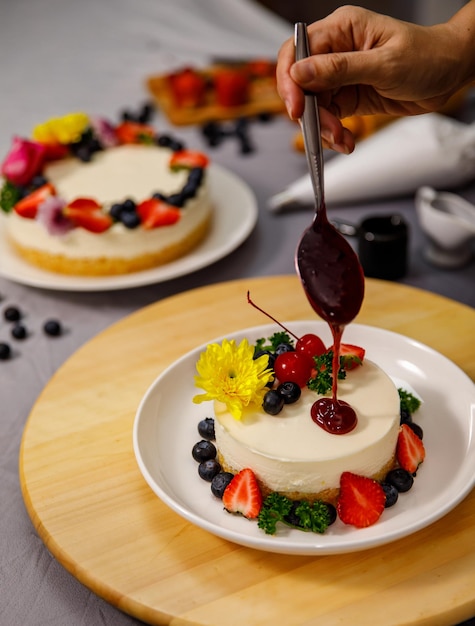 Verter salsa de arándanos en un delicioso pastel de queso dulce decorado con bayas frescas y flores en un plato de madera, para un cumpleaños, día de San Valentín y celebración