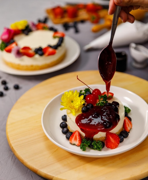 Verter salsa de arándanos en un delicioso pastel de queso dulce decorado con bayas frescas y flores en un plato de madera, para un cumpleaños, día de San Valentín y celebración