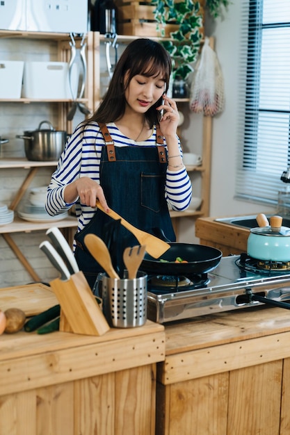 vertical de uma jovem esposa asiática está falando no celular no lazer enquanto frita legumes para o almoço no fogão a gás na cozinha em casa.