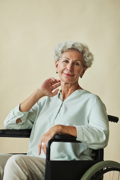 Vertical de elegante mujer senior mirando a la cámara en silla de ruedas contra un fondo neutro