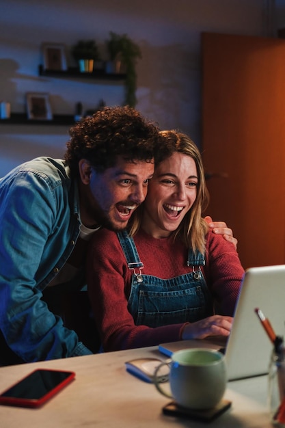 Vertical Feliz joven pareja caucásica sonriendo y divirtiéndose viendo videos en las redes sociales juntos usando una computadora portátil con conexión a Internet sentados en casa a altas horas de la noche