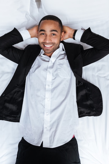 Vertical del hombre africano sonriente en traje acostado en la cama y mirando a la cámara