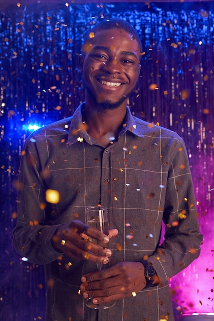 Vertical del joven afroamericano sosteniendo una copa de champán y sonriendo a la cámara mientras disfruta de la fiesta en la discoteca