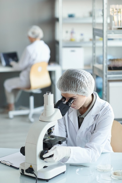 Vertical de la joven científica mirando al microscopio mientras estudia muestras de plantas en el laboratorio de biotecnología