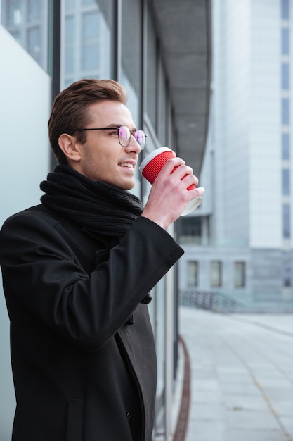 Vertical del joven empresario de gafas y ropa de abrigo de pie hacia los lados y bebiendo café al aire libre